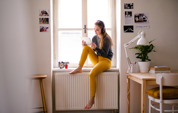 A young happy college female student sitting on window sill at home, using tablet and headphones when studying.