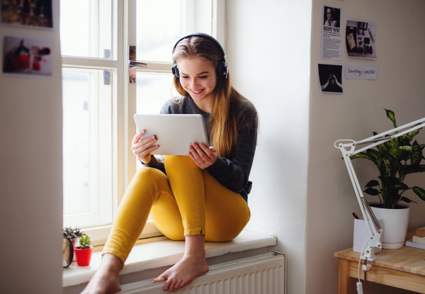 A young happy college female student sitting on window sill at home, using tablet and headphones when studying.