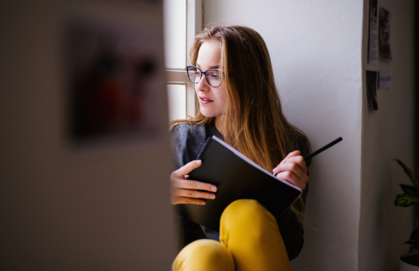 A young happy college female student with an exercise book sitting on window sill at home, studying.