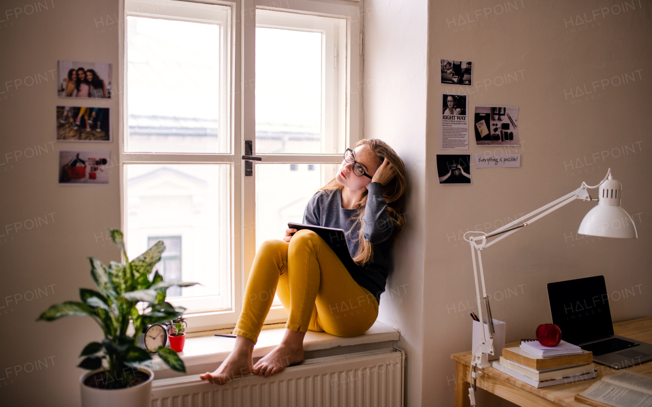 A young happy college female student with an exercise book sitting on window sill at home, studying.