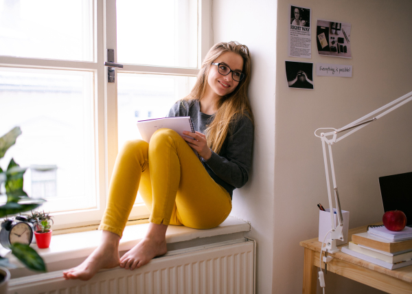 A young happy college female student with an exercise book sitting on window sill at home, studying.