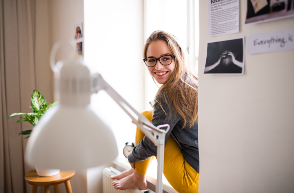 A young happy college female student sitting on window sill at home, resting after studying.