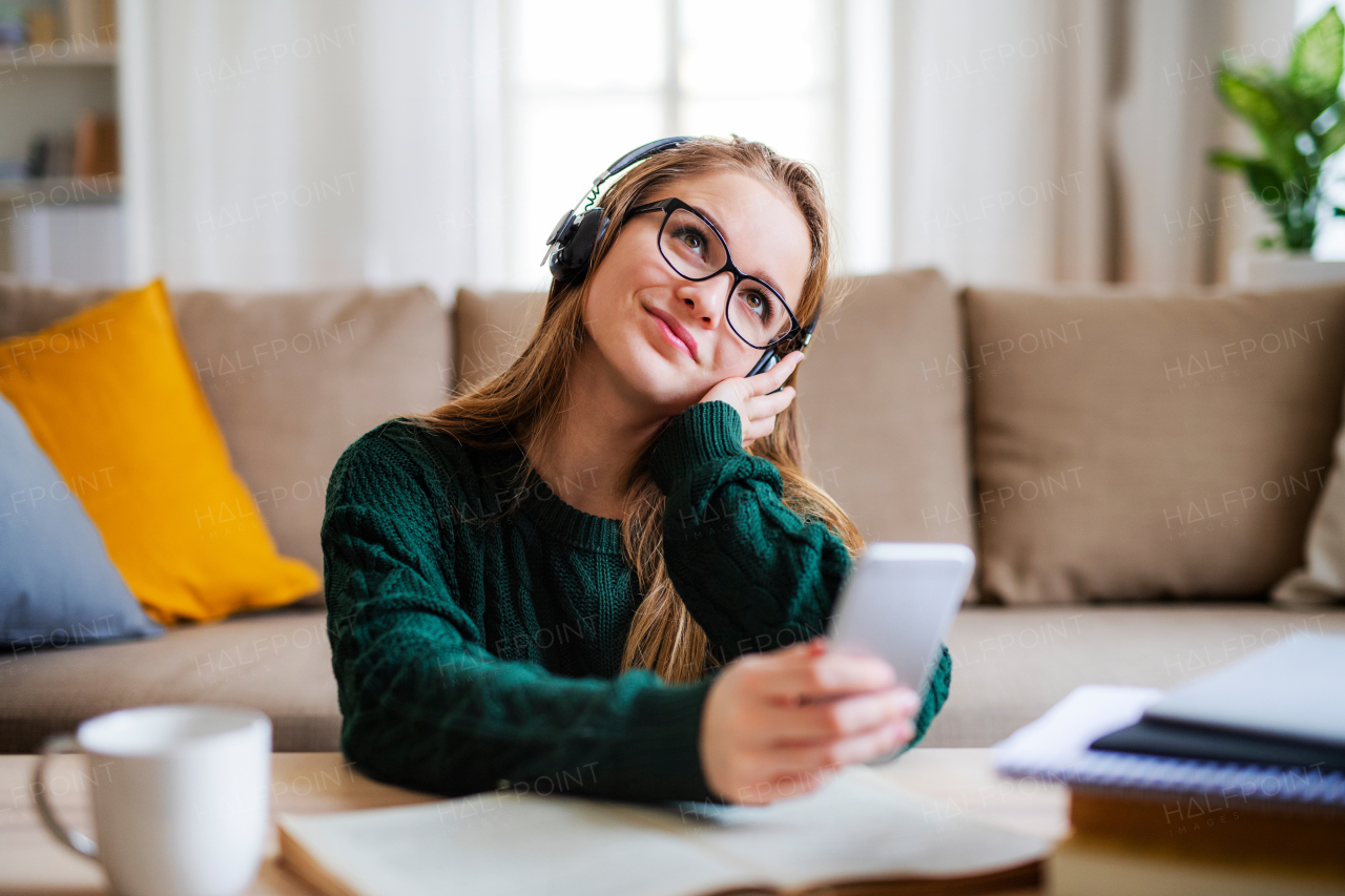 A young happy college female student sitting at the table at home, using headphones when studying.