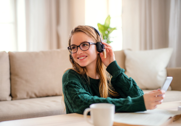 A young happy college female student sitting at the table at home, using headphones when studying.