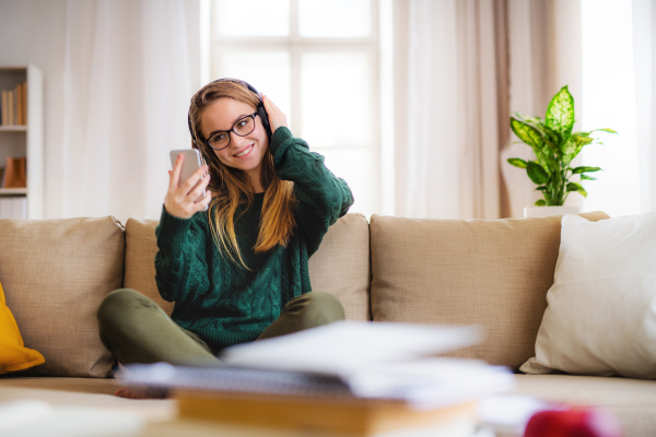 A young happy college female student sitting on sofa at home, using headphones when studying.