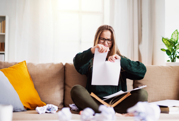 An unhappy, sad and frustrated female student sitting on sofa, tearing paper when studying.