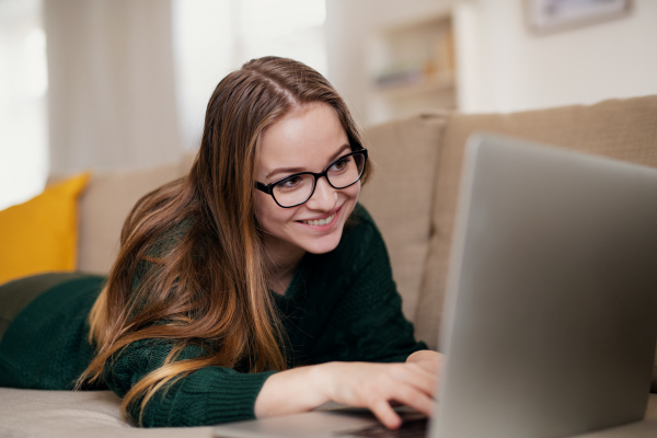 A young happy college female student lying down on sofa at home, using laptop when studying.