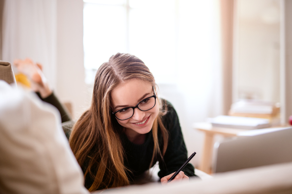 A young happy college female student lying down on sofa at home, writing and studying.