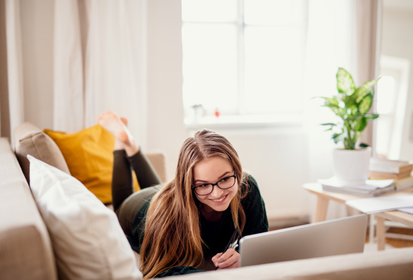 A young happy college female student lying down on sofa at home, using laptop when studying.