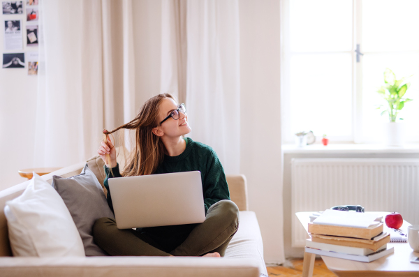 A young happy college female student sitting on sofa at home, using laptop when studying.