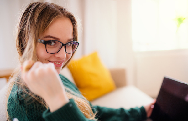 A young happy college female student sitting on sofa at home, using laptop when studying.