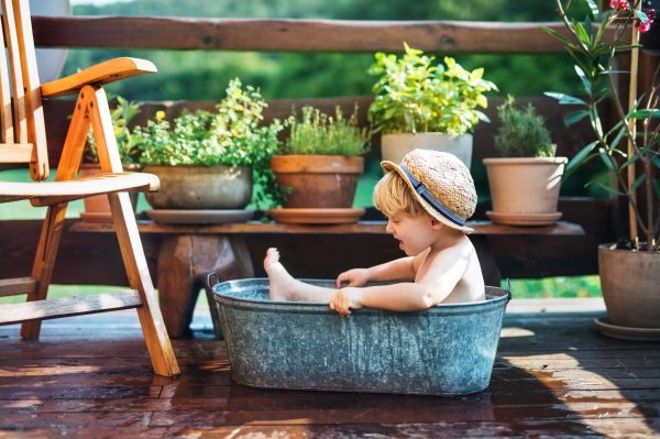 Happy small boy with a hat in bath tub outdoors in garden in summer, playing in water.