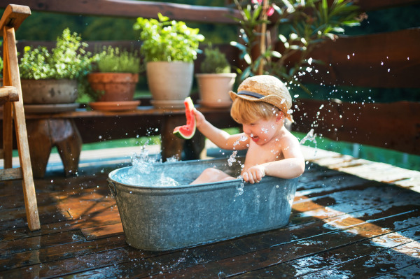 Happy small boy with a hat in bath tub outdoors in garden in summer, eating watermelon.