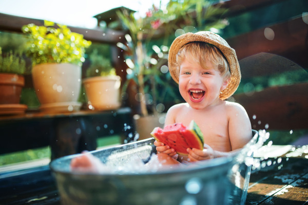 Happy small boy with a hat in bath tub outdoors in garden in summer, eating watermelon.