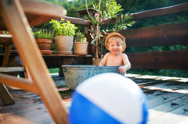 Happy small boy with a ball in bath tub outdoors in garden in summer, playing in water.