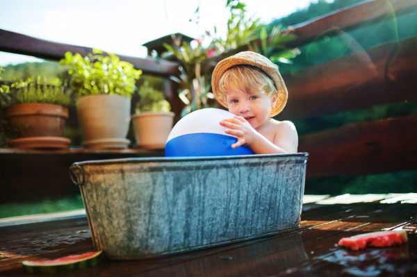 Happy small boy with a ball in bath tub outdoors in garden in summer, playing in water.