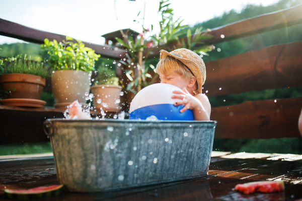 Happy small boy with a ball in bath tub outdoors in garden in summer, playing in water.