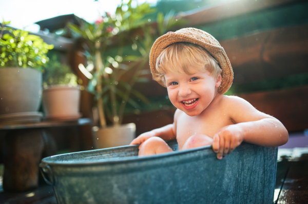 Happy small boy with a hat in bath tub outdoors in garden in summer, playing in water.