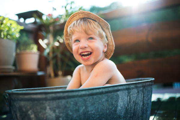 Happy small boy with a hat in bath tub outdoors in garden in summer, playing in water.