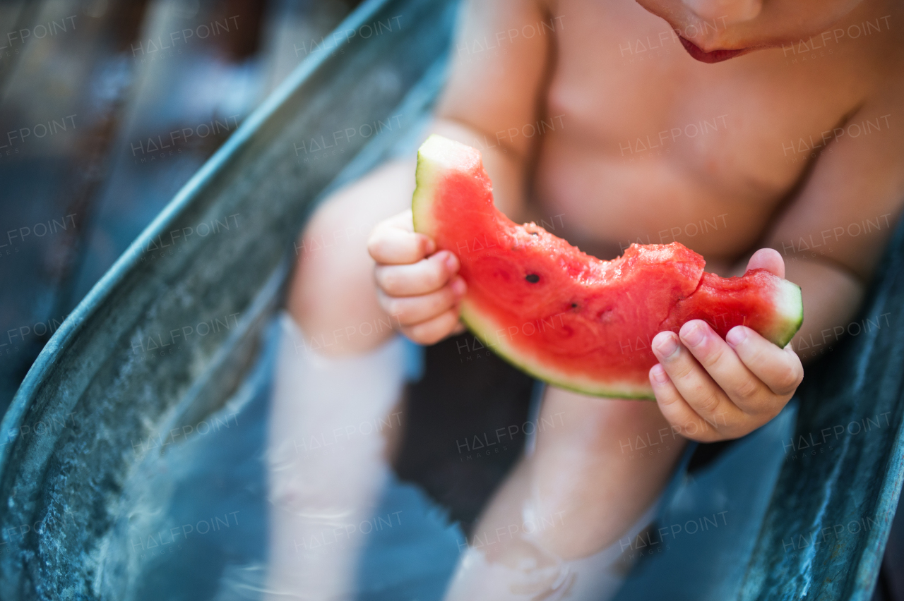 A midsection of unrecognizable small boy in bath outdoors in garden in summer, eating watermelon.