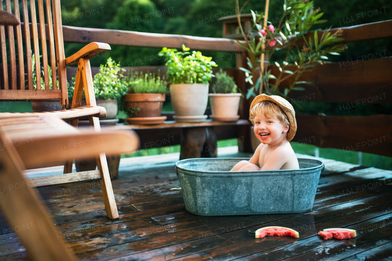 Happy small boy with a hat in bath tub and watermelon outdoors in garden in summer.