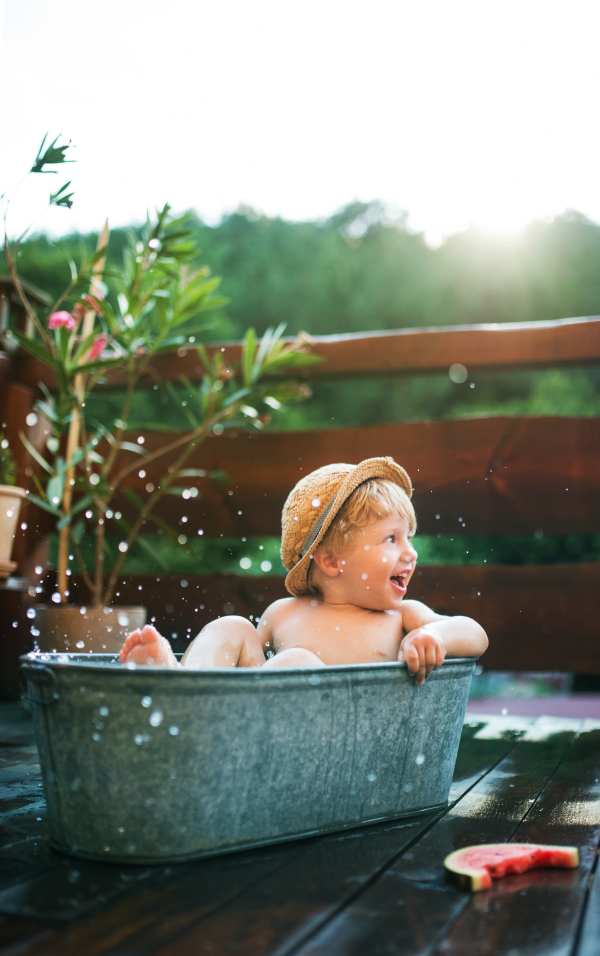 Happy small boy with a hat in bath tub outdoors in garden in summer, eating watermelon.