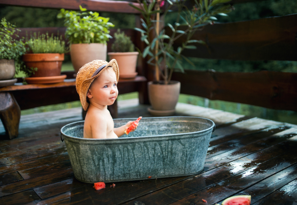 Happy small boy with a hat in bath tub outdoors in garden in summer, eating watermelon.