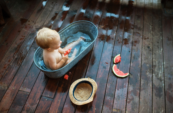 A top view of happy small girl in bath tub outdoors in garden in summer, eating watermelon.