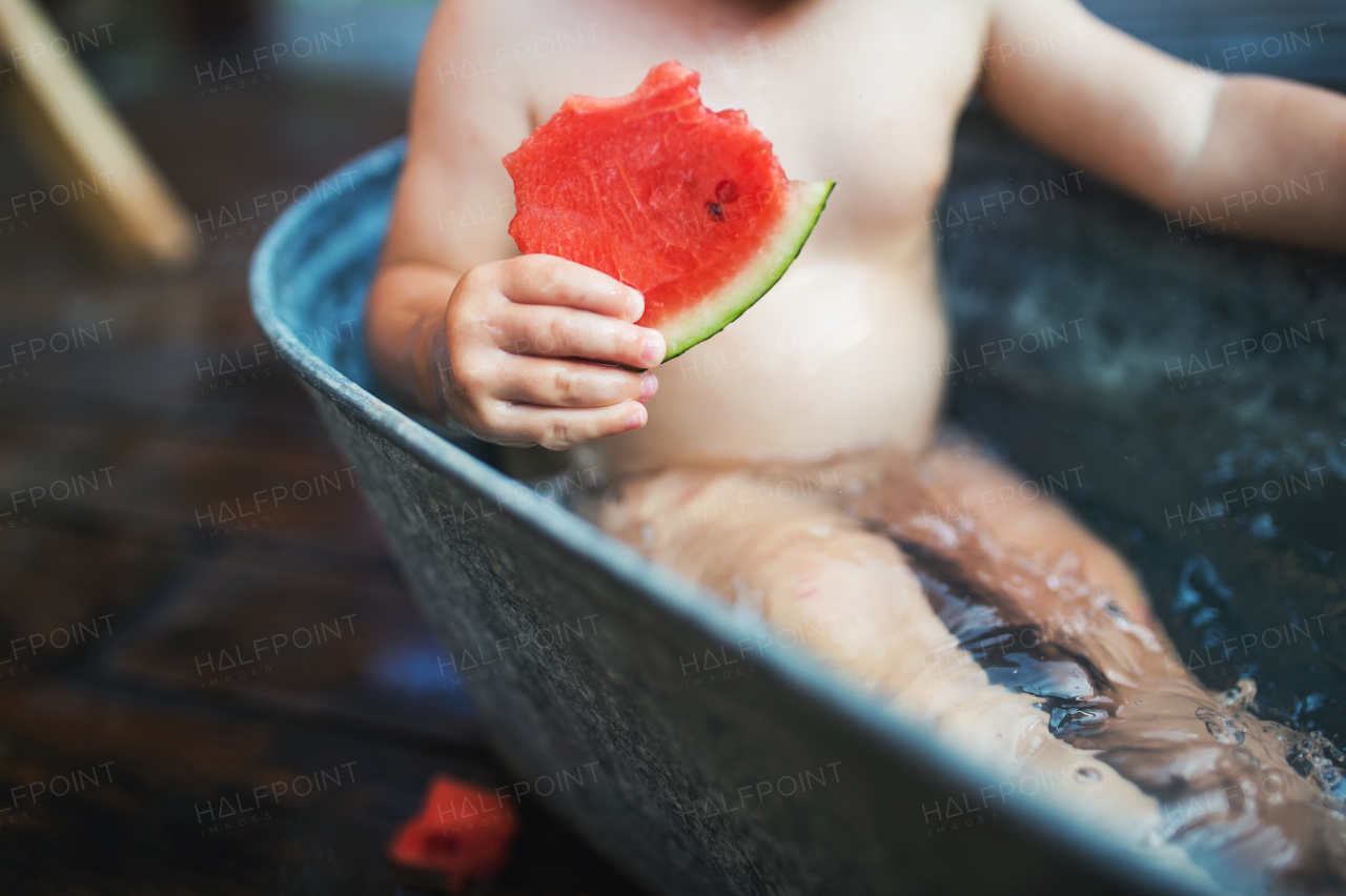 A midsection of unrecognizable small boy or girl in bath outdoors in garden in summer, eating watermelon.