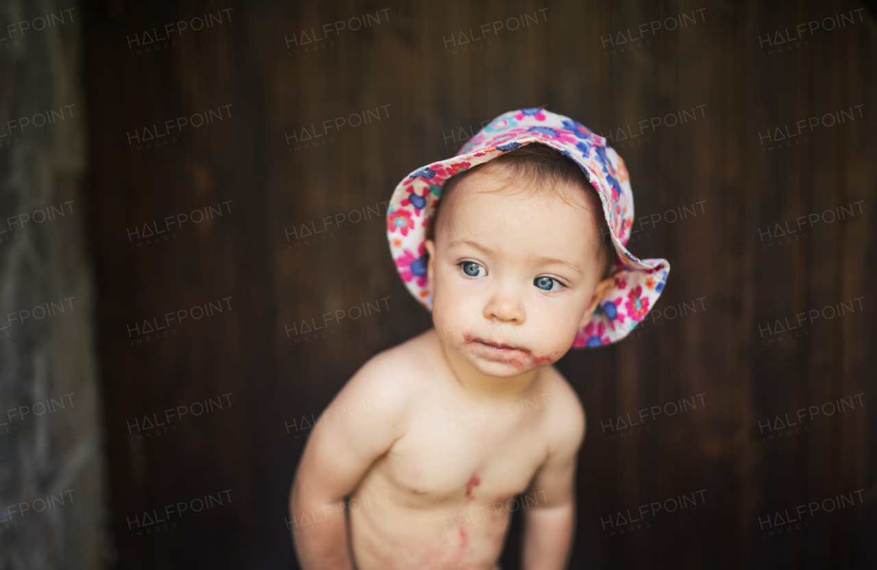 A front view of small girl standing against dark wooden background on a patio in summer. Copy space.