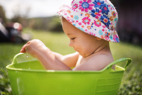 Small girl with a hat sitting in plastic bucket outdoors in garden in summer, playing in water.