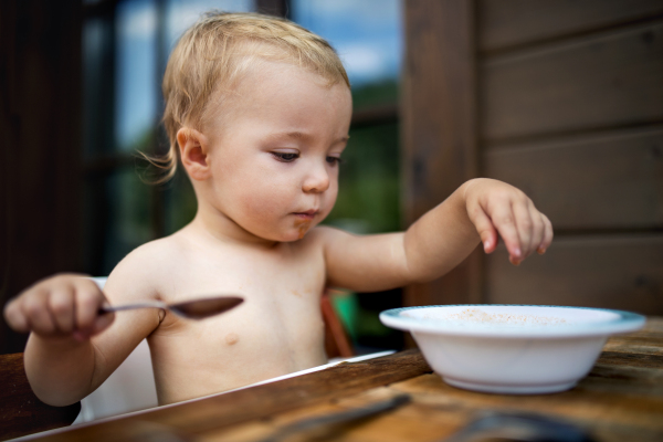 Cute topless small girl standing by a table on a patio in summer, eating soup.