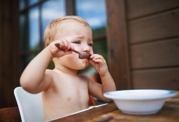 Cute topless small girl standing by a table on a patio in summer, eating soup.