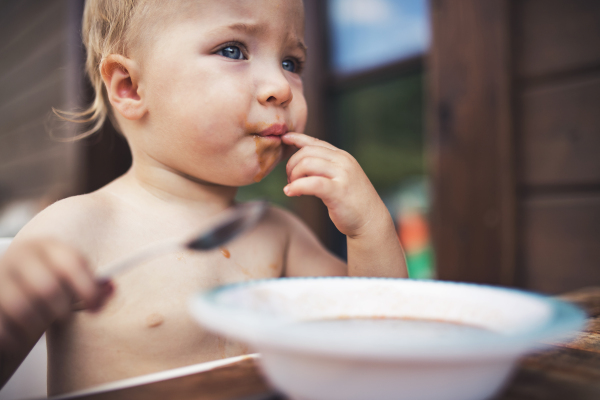 Cute topless small girl standing by a table on a patio in summer, eating soup.