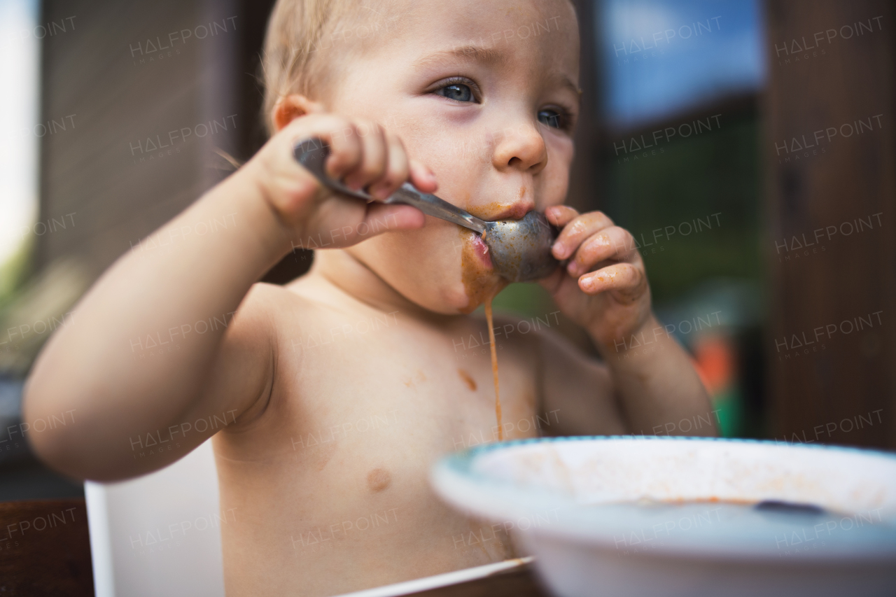 Cute topless small girl standing by a table on a patio in summer, eating soup.