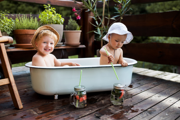 Small children with a drink sitting in bath outdoors in garden in summer, playing in water.