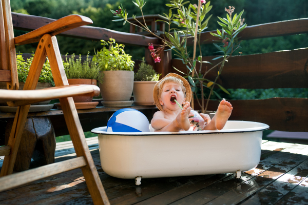 Happy small boy with a hat in bath tub outdoors in garden in summer, playing in water.