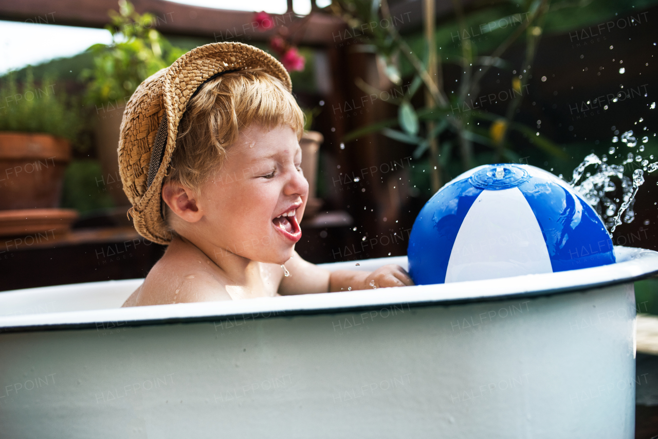 Happy small boy with a ball in bath tub outdoors in garden in summer, playing in water.