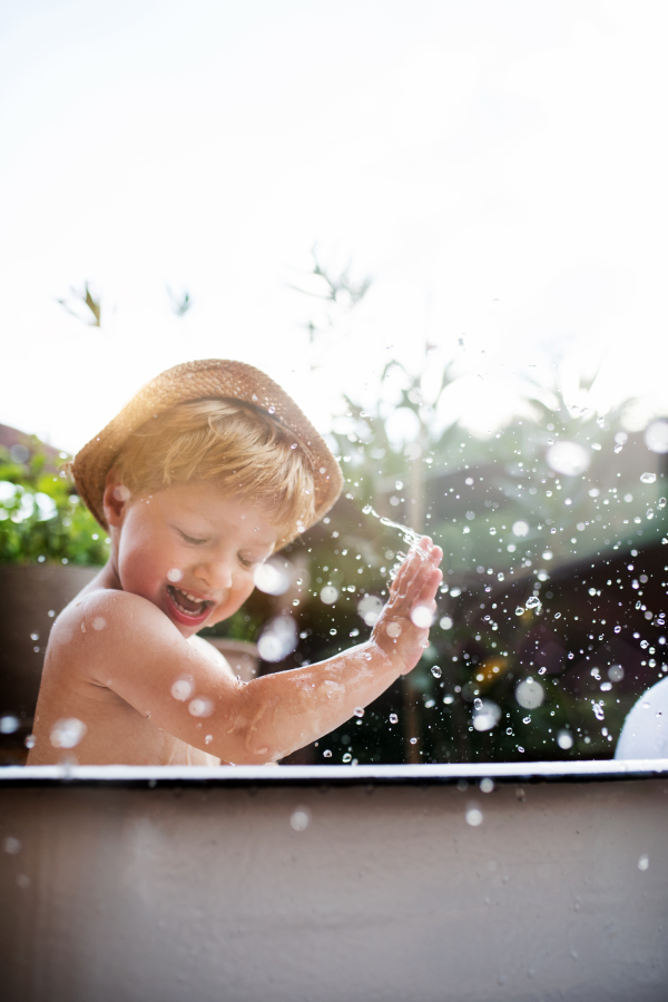 Happy small boy with a hat in bath tub outdoors in garden in summer, playing in water.