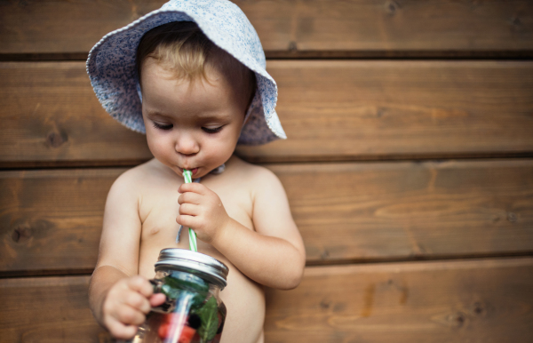 A front view of small girl with a drink standing against dark wooden background on a patio in summer. Copy space.