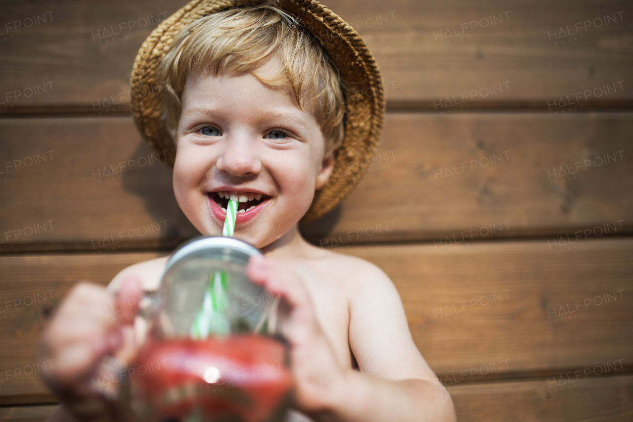 A front view of small boy with a drink standing against dark wooden background on a patio in summer. Copy space.