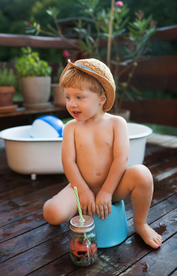 Small boy with a hat and drink in garden in summer, sitting on a potty.