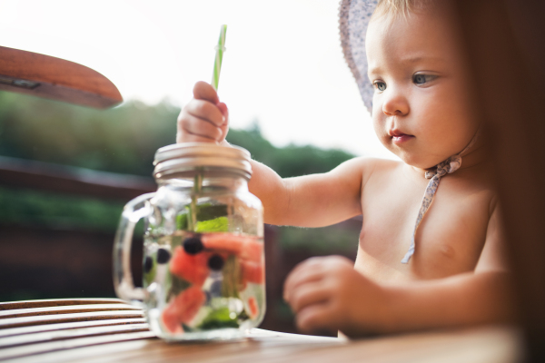 A small girl with a drink wth fruit standing by a table on a patio in summer.