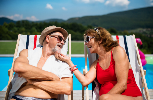 Cheerful senior couple in love sitting by swimming pool outdoors in backyard.