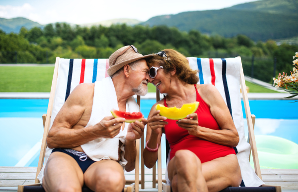 Cheerful senior couple in love sitting by swimming pool outdoors in backyard, talking.