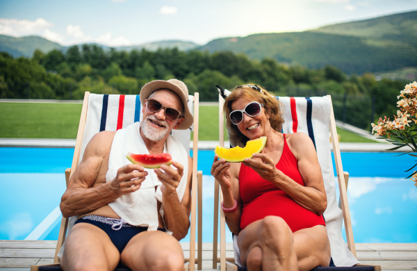 Cheerful senior couple sitting by swimming pool outdoors in backyard, looking at camera.