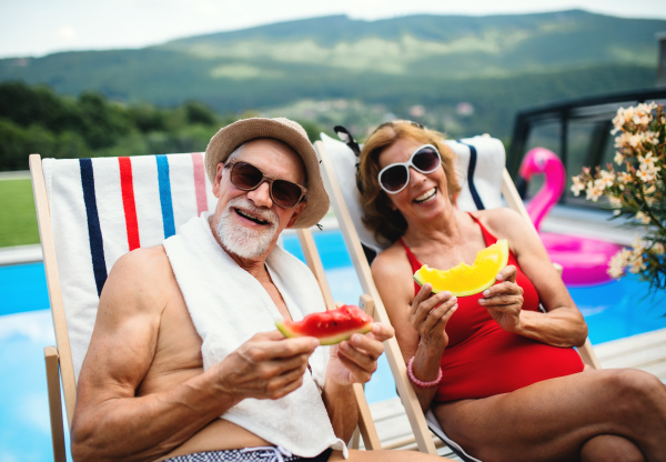 Cheerful senior couple sitting by swimming pool outdoors in backyard, looking at camera.