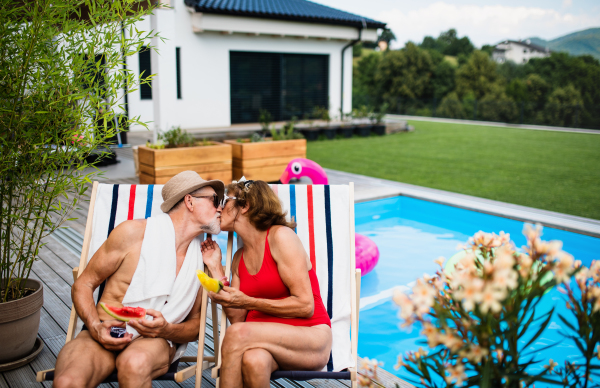 Cheerful senior couple in love sitting by swimming pool outdoors in backyard, kissing.