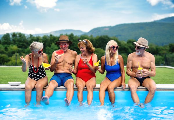 Group of cheerful seniors sitting by swimming pool outdoors in backyard, talking.