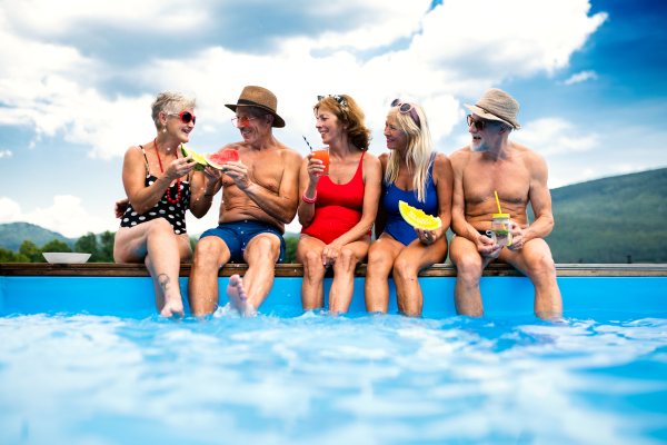 Group of cheerful seniors sitting by swimming pool outdoors in backyard, talking.